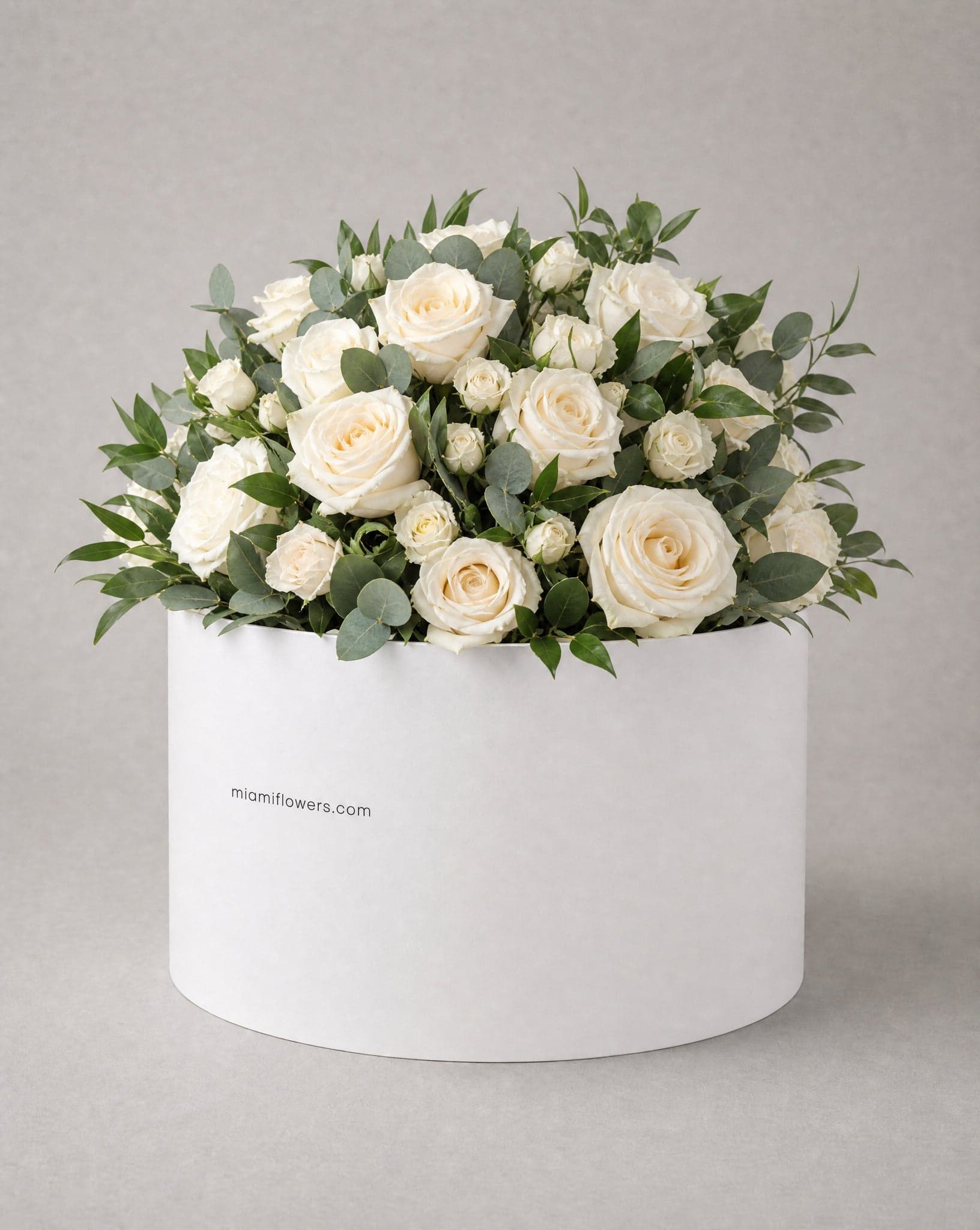 Cream roses and dark green foliage in a tall matte white oval box printed with miamiflowers.com