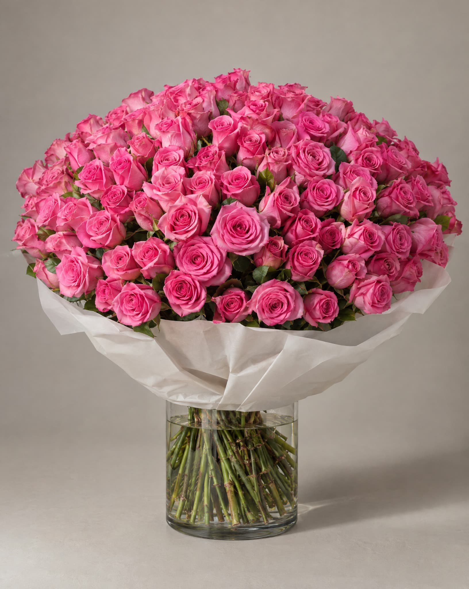 One hundred bright pink roses in a domed bouquet wrapped in white paper in a clear glass vase