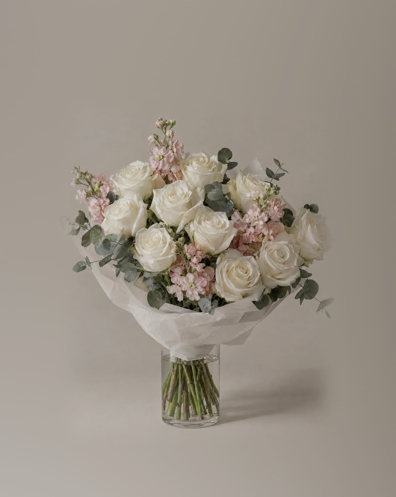 A dozen white roses with pale pink stock and eucalyptus, wrapped in white tissue in a clear glass vase