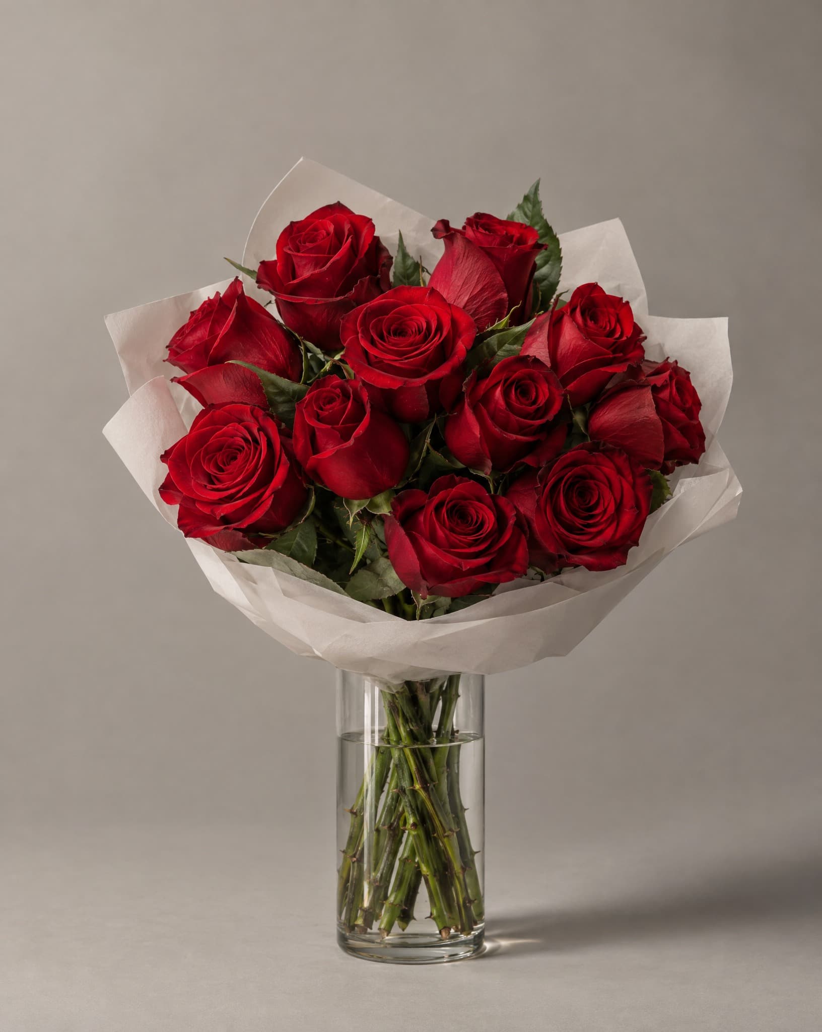 A dozen deep red roses hand-wrapped in white tissue, standing in a clear glass vase