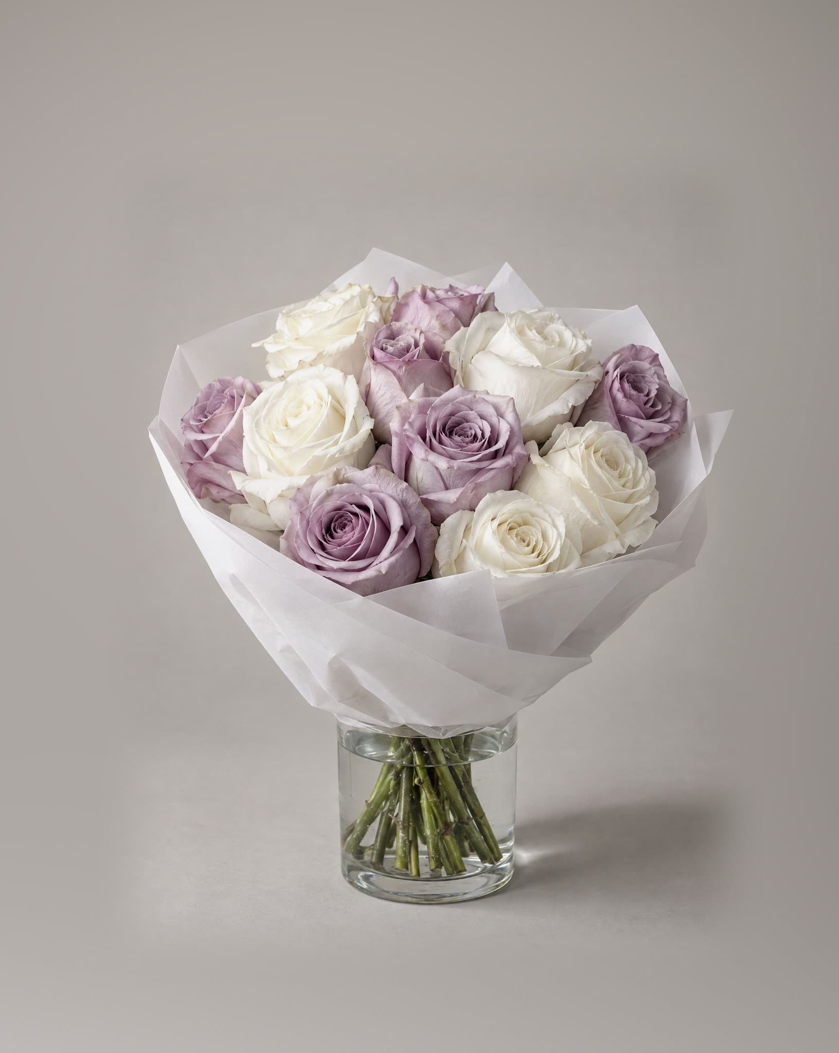 A dozen lavender and ivory roses wrapped in white paper in a clear glass vase