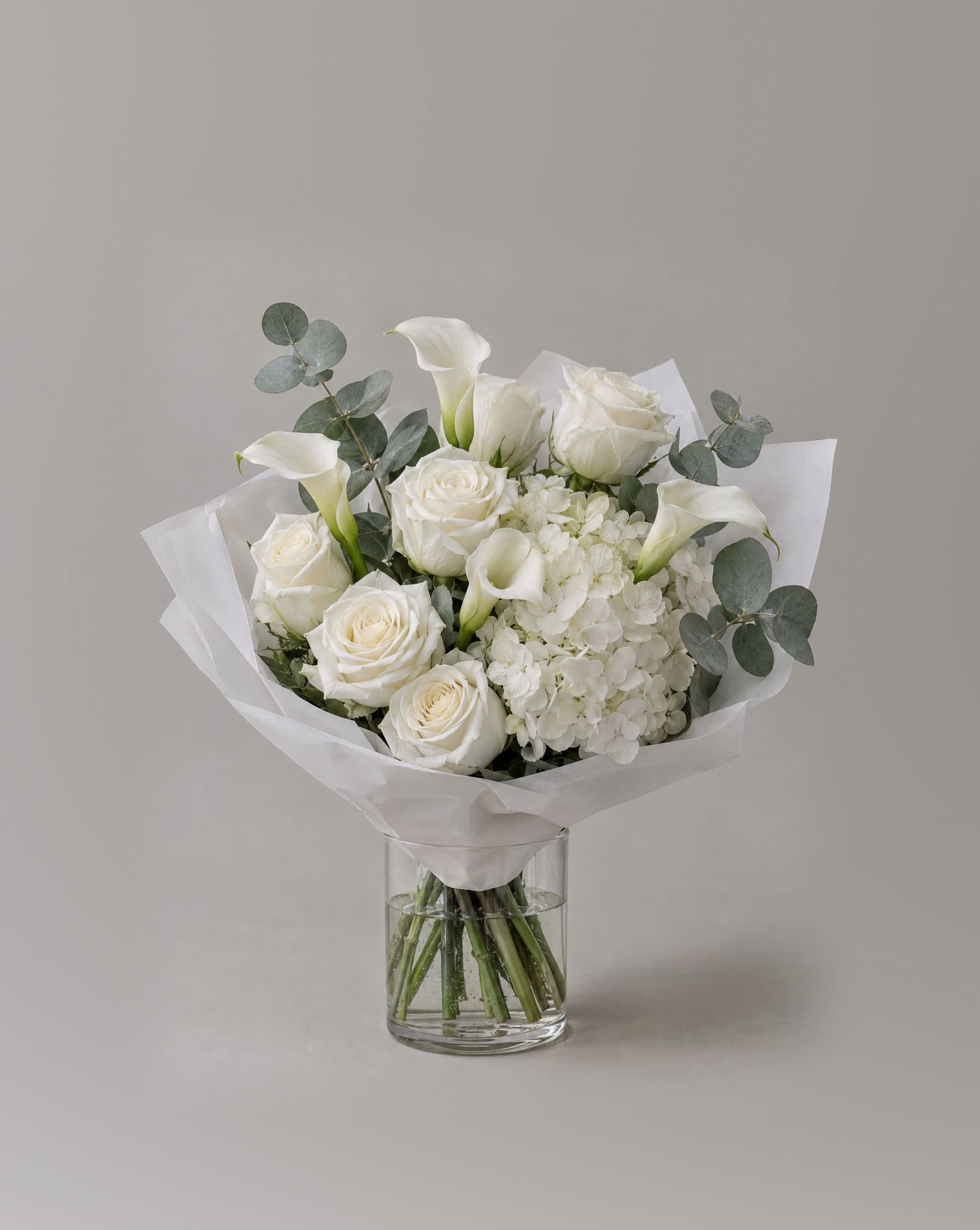 Standard bouquet of white hydrangeas, white roses and white calla lilies with eucalyptus in a clear glass vase