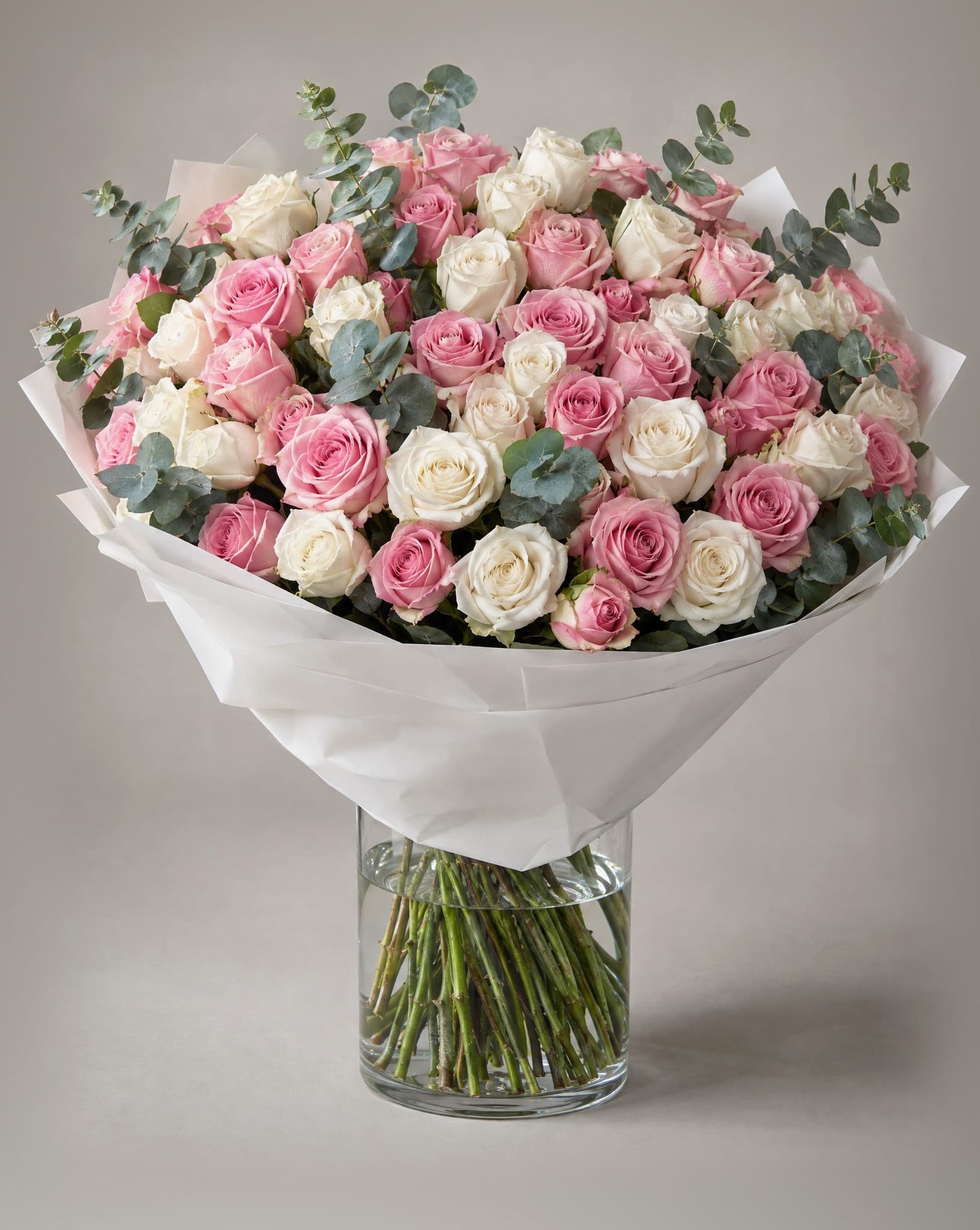 One hundred pink and white roses with eucalyptus wrapped in white paper in a clear glass vase