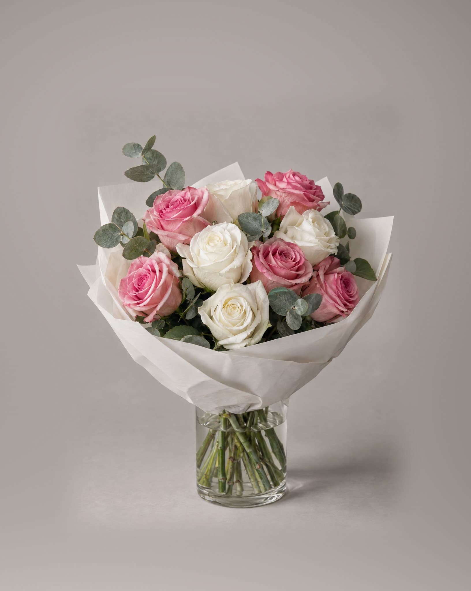 A dozen pink and white roses with eucalyptus wrapped in white paper in a clear glass vase