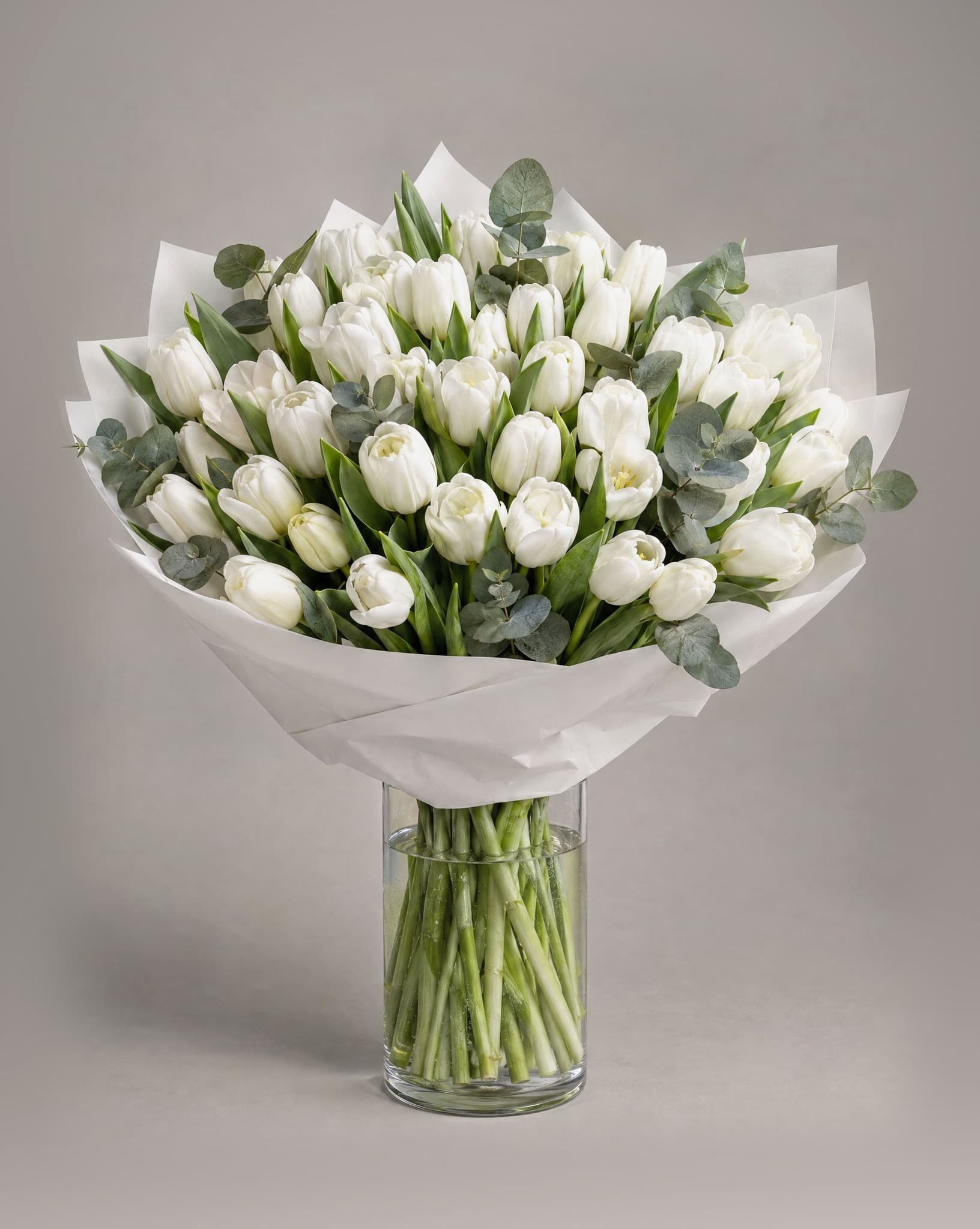 Grand bouquet of white tulips with eucalyptus wrapped in white paper in a clear glass vase