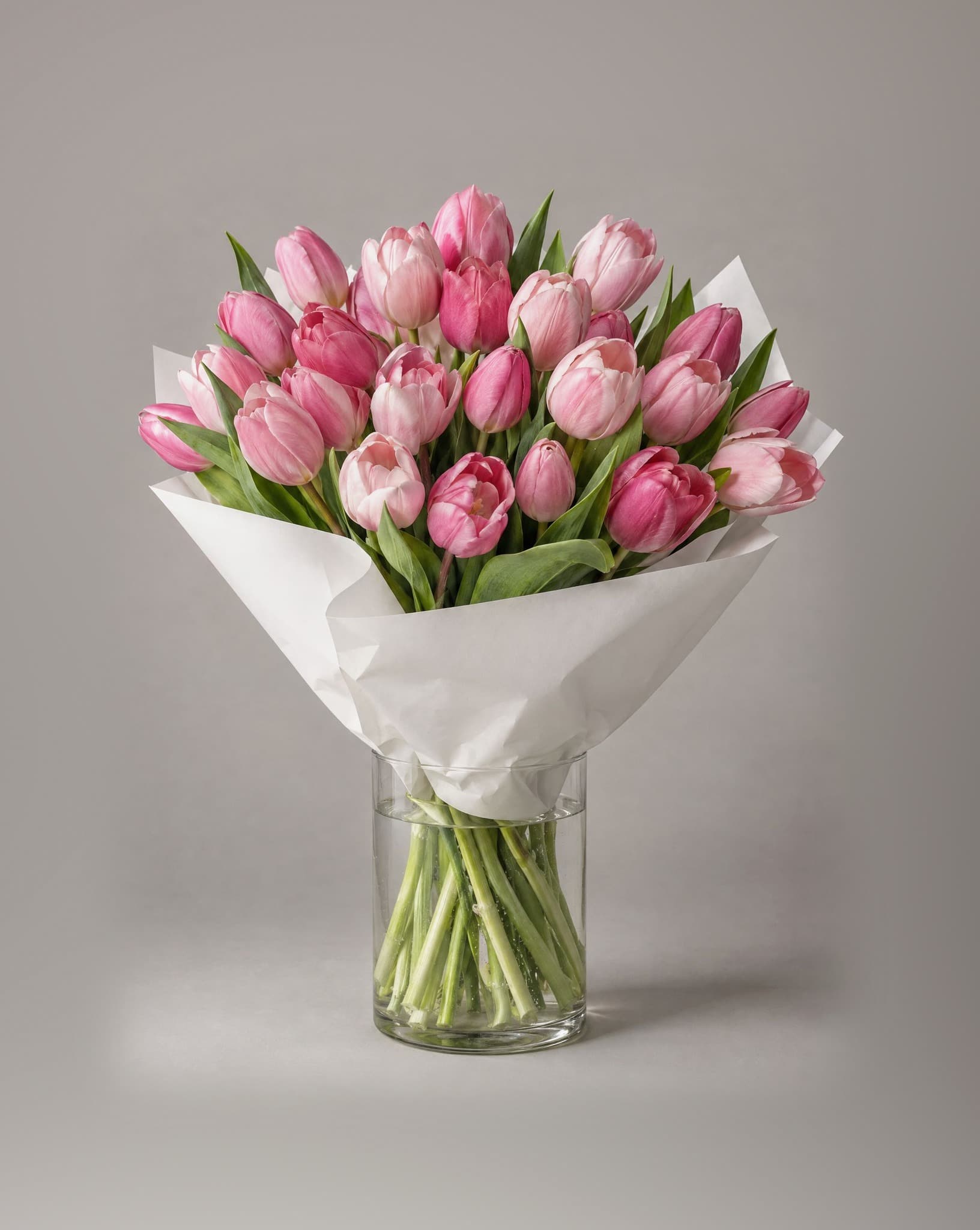 Standard bouquet of pink tulips in shades from blush to rose wrapped in white paper in a clear glass vase